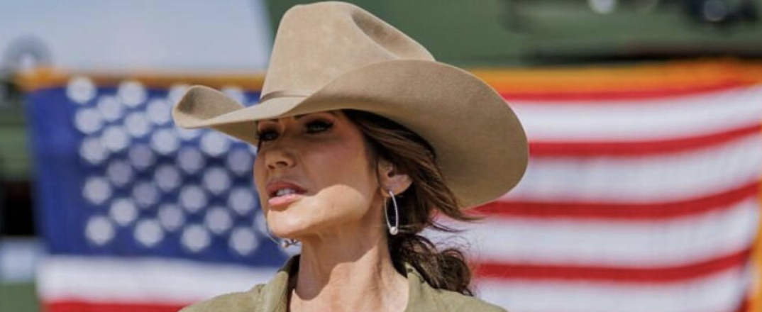Kristi Noem in cowboy hat standing in front of large American flag